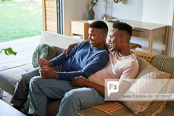 Happy young gay male couple using digital tablet on living room sofa