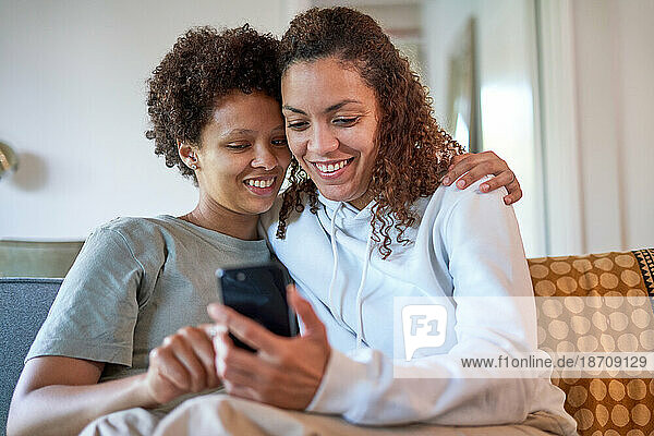 Happy lesbian couple hugging  using smart phone on sofa at home