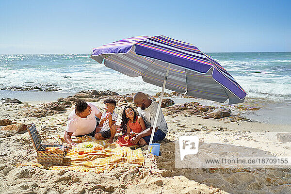 Gay male couple and kids eating lunch under umbrella on sunny beach