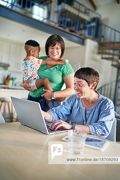 Happy lesbian couple with son using laptop in dining room at home