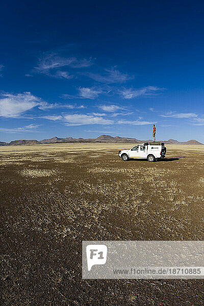 Male standing on top of expedition truck in the middle of African desert. Wide open space and blue sky.