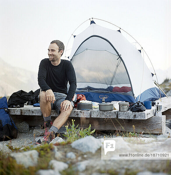 A smiling man prepares a meal beside his tent.