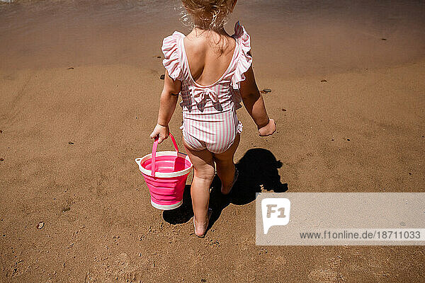 Child on sandy beach walking away with pink bucket on summer vac