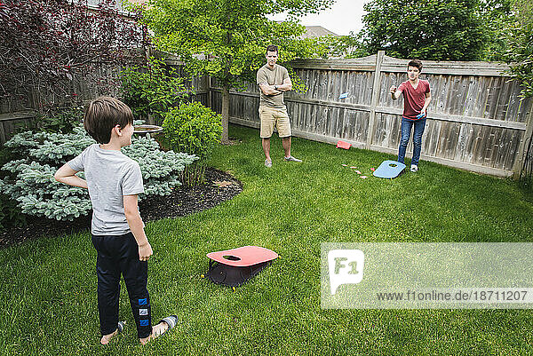 Two boys playing corn hole game in backyard with dad watching them.
