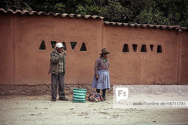 Native Peruvians stand in front of mud wall waiting for bus