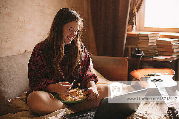 Teenage girl eating chips while sitting on bed with laptop