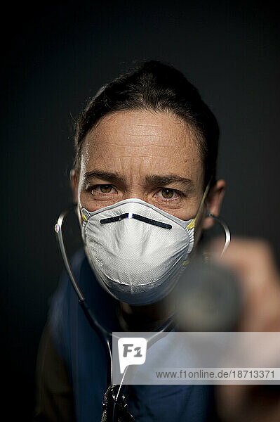 Studio portrait of 42 year old caucasian female registered nurse wearing a N95 mask and pointing a stethoscope at the camera.