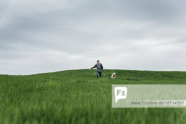 Teenage boy running across a grassy field with his dog on cloudy day.