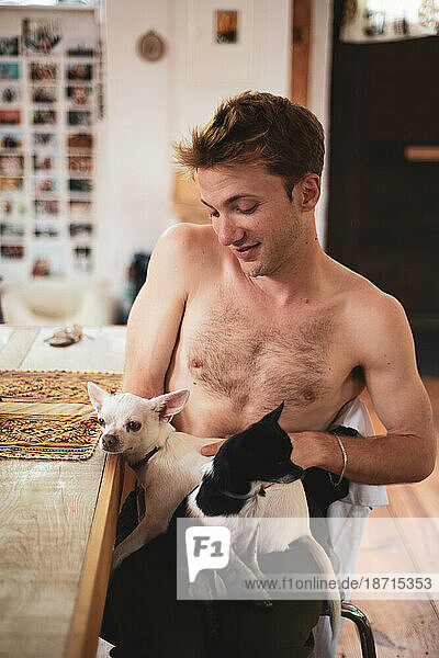shirtless man smiles and holds two small chihuahua dogs at home