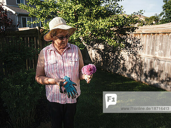Older woman in hat looking at bunch of freshly cut flowers in a yard.