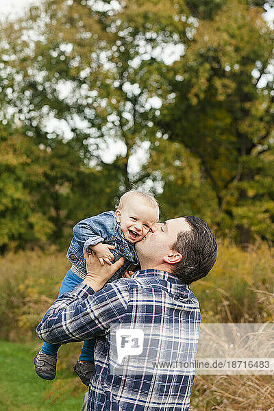 A happy father raises his laughing baby boy up to for a kiss in a park