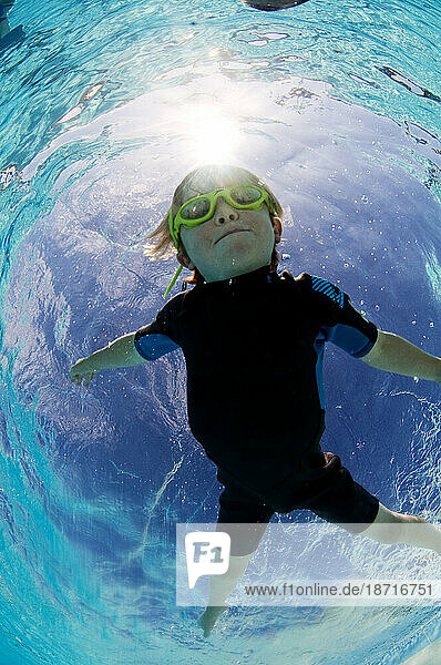 Underwater view of a young boy  wearing goggles  floating in a swimming pool.