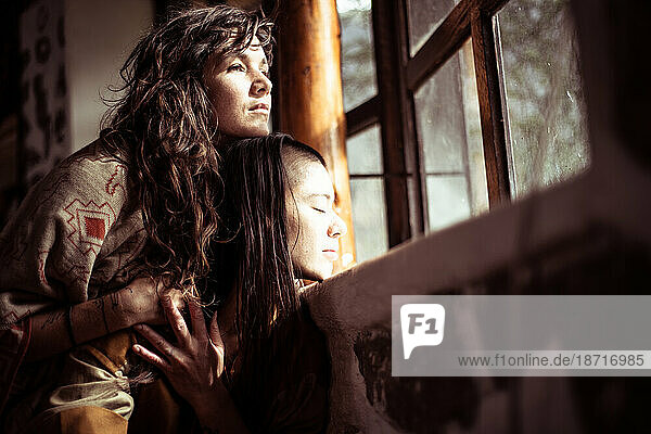 Same sex lesbian couple cuddle and look out window with light on face