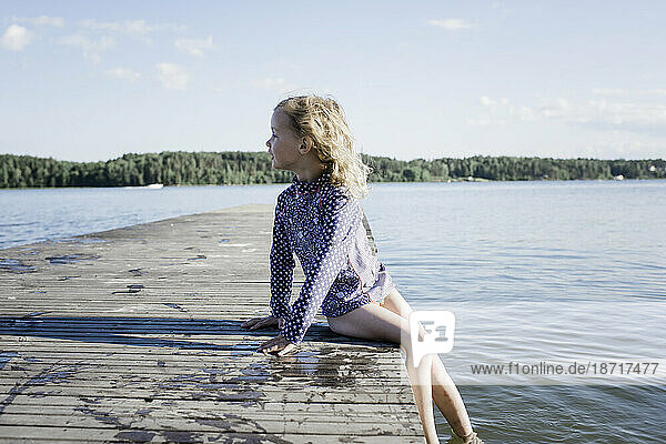 young blonde girl sat on a pier looking out to see