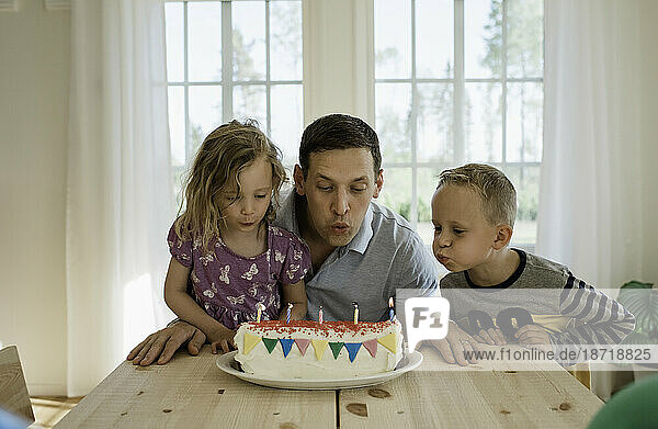 a father with his daughter & son blowing candles on a birthday cake