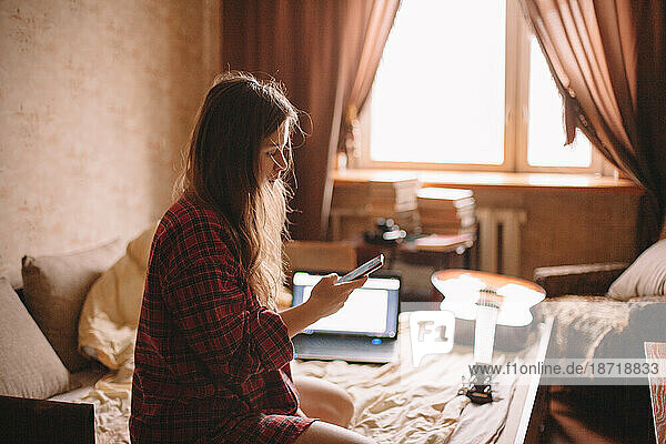 Young woman using smart phone while sitting on bed at home