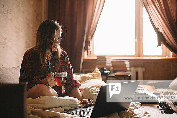 Young woman holding cup of tea while using laptop in bed
