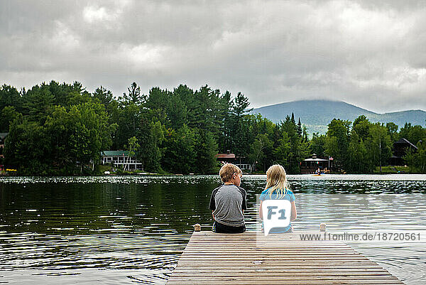 view from behind boy and girl sitting at edge of dock on lake