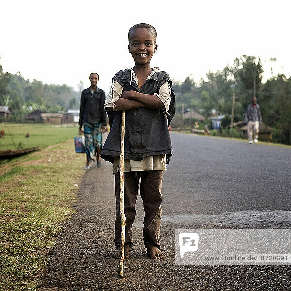 A boy with a walking stick poses on the edge of a paved road with villagers walking the road behind him.