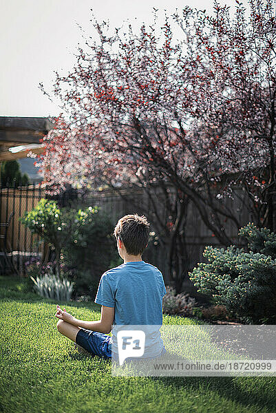 Back view of tween boy sitting on the grass in a garden meditating.