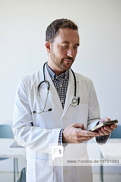 Male doctor surfing net through smart phone while standing in clinic