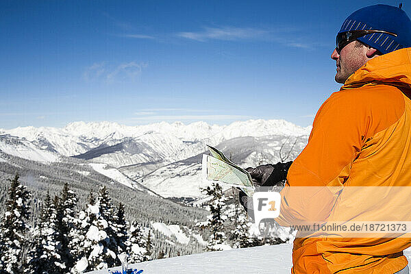 A young man wearing orange looks at a map from a high vantage point above a valley below.