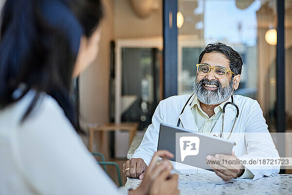 Happy doctor holding tablet PC while discussing with patient in clinic