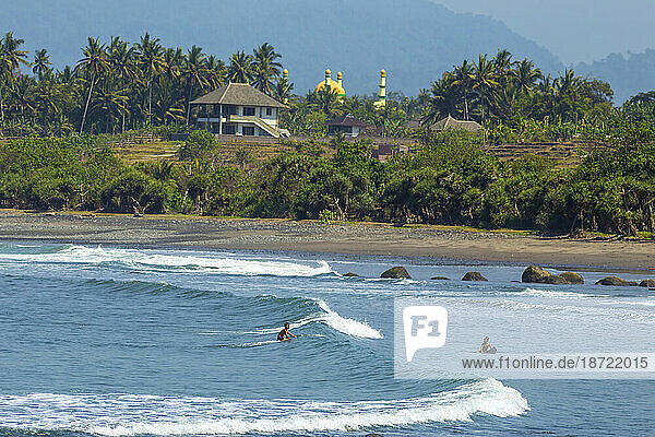 Balinese children surf a wave at Medewi beach with with a mosque at background. . Bali. Indonesia.