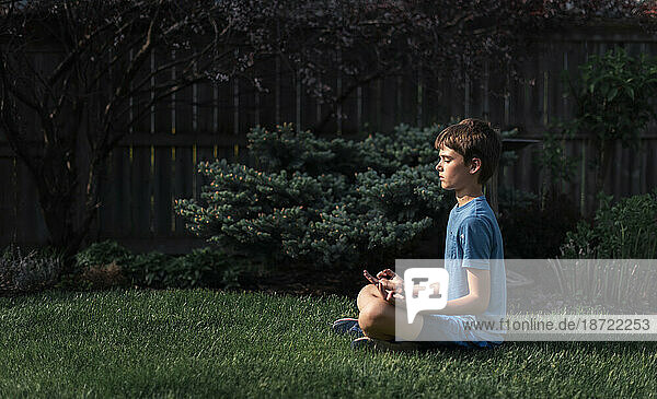 Side view of tween boy sitting on the grass in a garden meditating.