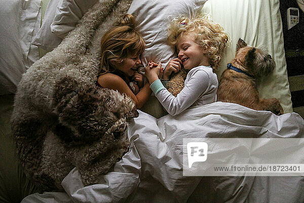 two sisters and dogs laying in bed holding hands giggling morning time