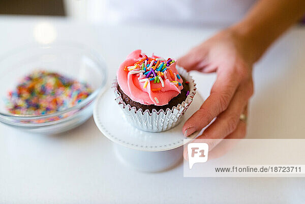 A woman's hand holding a decorated cupcake with a bowl of sprinkles