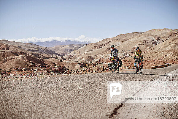 Two bike packers ride along a road with desert mountains behind them