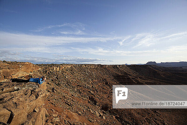 A woman lays on her belly on the ground  looking over the edge of a rocky cliff . Canyonlands National Park in Utah