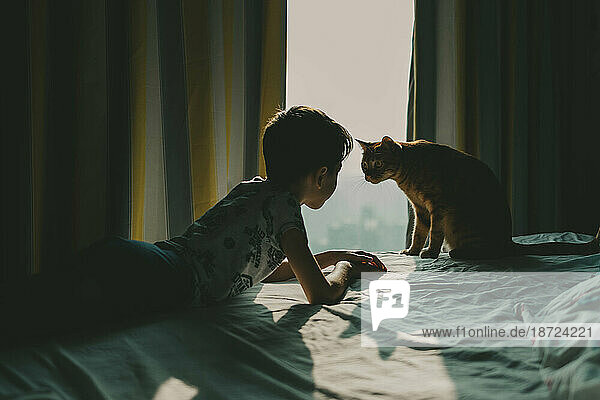 The boy and the cat lie on the bed and look at each other