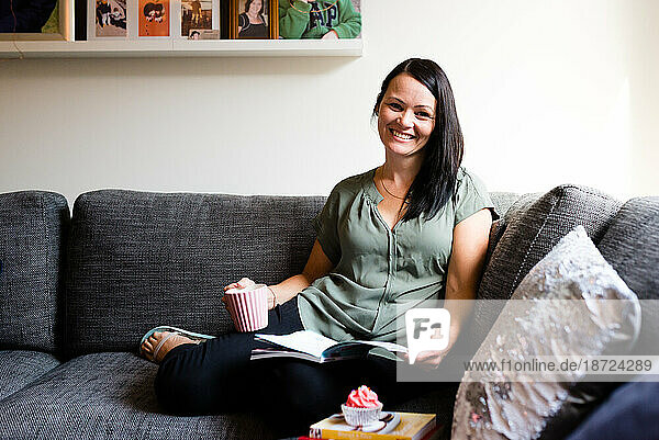 A woman smiling and reading on the sofa with a cupcake