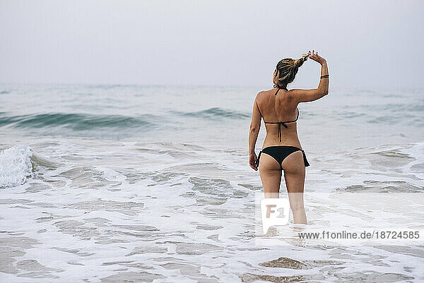 Back view of woman with black bikini arranging her hair on the beach.
