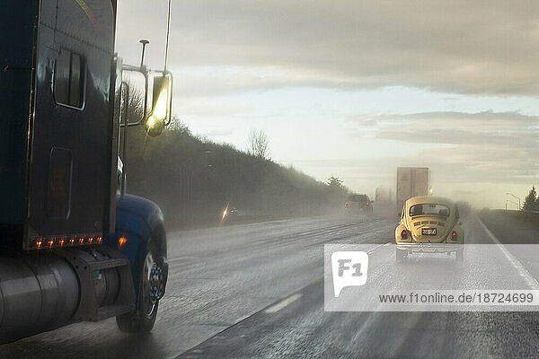 An old model yellow car drives on the rainy highway next to a commercial truck near Seattle  Washington.