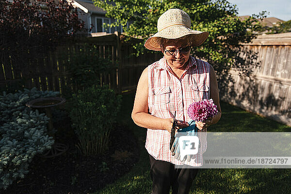 Older woman in hat holding bunch of freshly cut flowers in a yard.