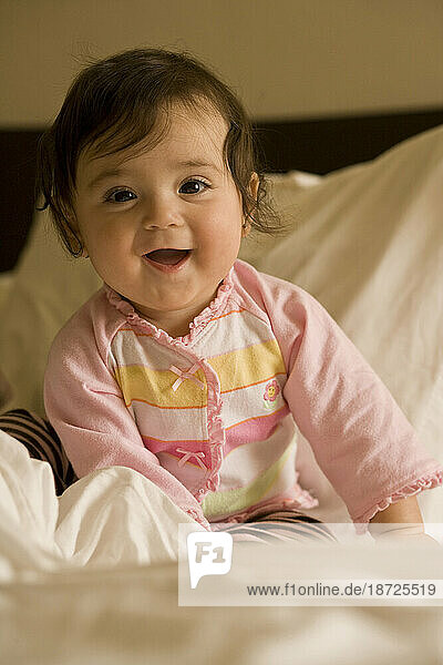 A happy baby girl sits on a bed surrounded by soft white sheets and smiles for the camera.