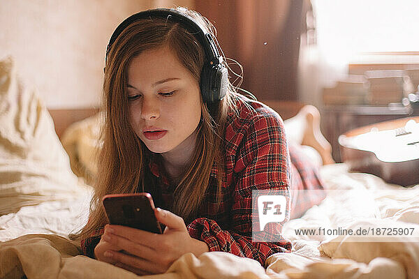 Teenage girl with headphones using smart phone lying on bed at home