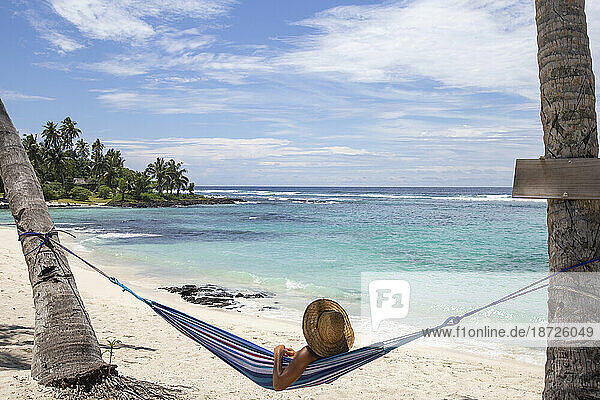 Man with straw hat  relaxing on blue hammock at sandy beach  Samoa