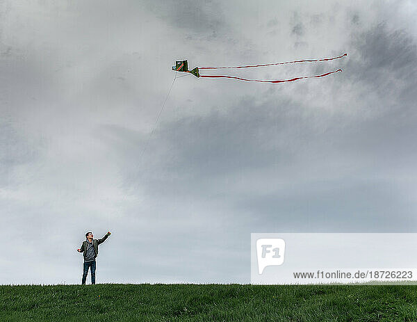 Teenage boy flying a kite on a grassy hill on a cloudy day.