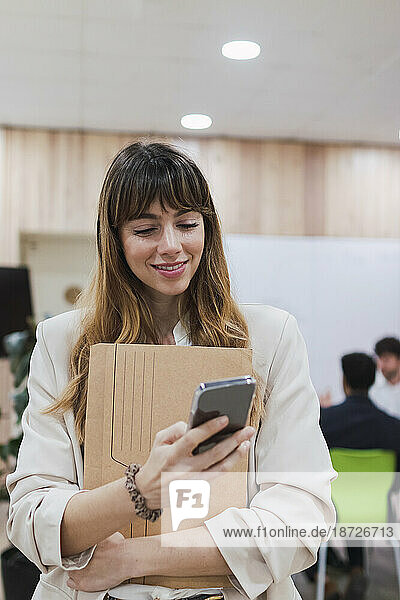 Smiling businesswoman holding folder and using mobile phone in office