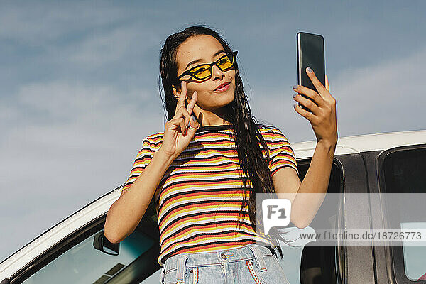 Smiling young woman taking selfie through smart phone leaning on car