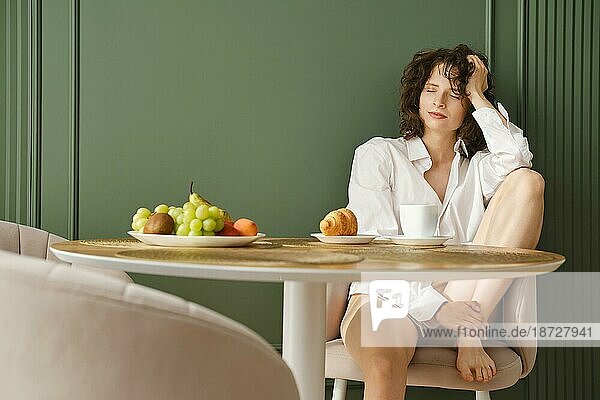 Verträumte Frau im weißen Hemd sitzt am Tisch in ihrer modernen Wohnung und genießt gesundes Obst und Morgenkaffee