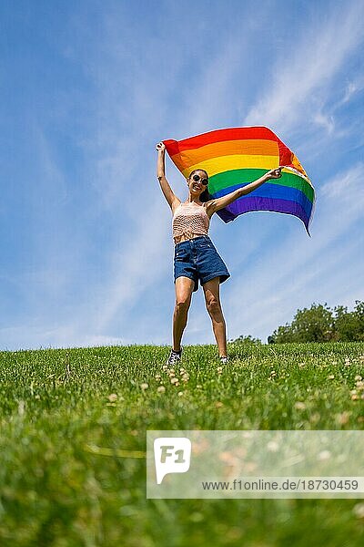 Caucasian brunette Frau mit einem Regenbogen lgbt Flagge auf dem Gras und blauen Himmel Hintergrund  winken die Flagge