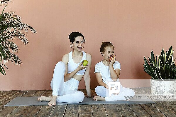Lächelnde Frau und kleines Mädchen essen Früchte nach dem Üben von Yoga auf der Matte im gemütlichen Wohnzimmer