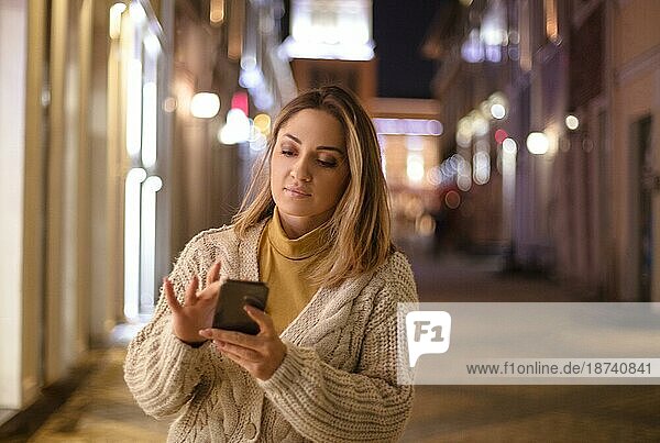 Junge attraktive Frau  die ihr Handy im Freien benutzt  eine Frau  die auf einer Straße in der Stadt spazieren geht und eine Nachricht auf einem modernen Smartphone tippt oder während eines Abendspaziergangs mit Freunden online chattet