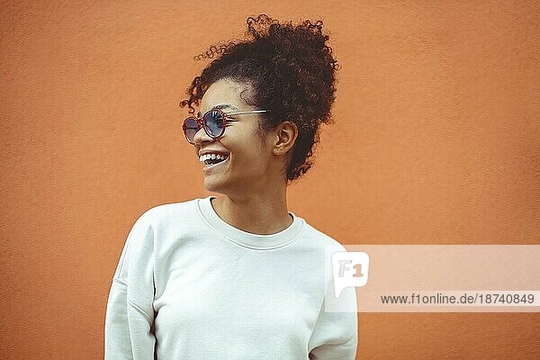 Junge Frau afrikanischer Abstammung mit stilvoller Sonnenbrille  mit lockigem Haar  das zu einem hohen Pferdeschwanz gebunden ist  schaut weg  während sie breit lächelt und gerade  perfekte Zähne zeigt  und vor einem orangefarbenen Wandhintergrund posiert