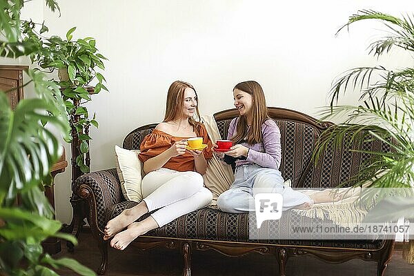 Junge  zufriedene Frauen sitzen auf einer gemütlichen  altmodischen Couch und plaudern beim Trinken von heißem Tee zwischen Blumentöpfen